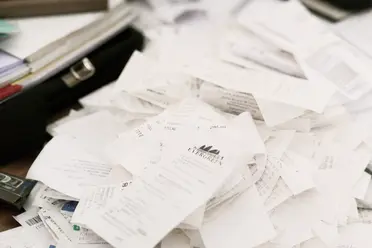 A messy pile of receipts and documents on a cluttered desk, with a stapler and some folders visible in the background.