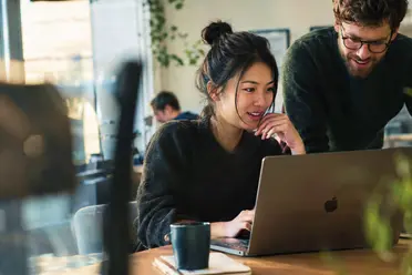 A woman and a man collaborate on a laptop in a cozy, sunlit workspace with plants in the background.