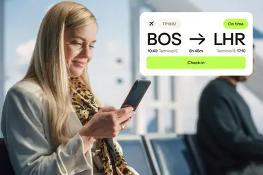 A woman at the airport checks her phone. Nearby, a departure sign shows flight TP1892 from Boston to London as on time.
