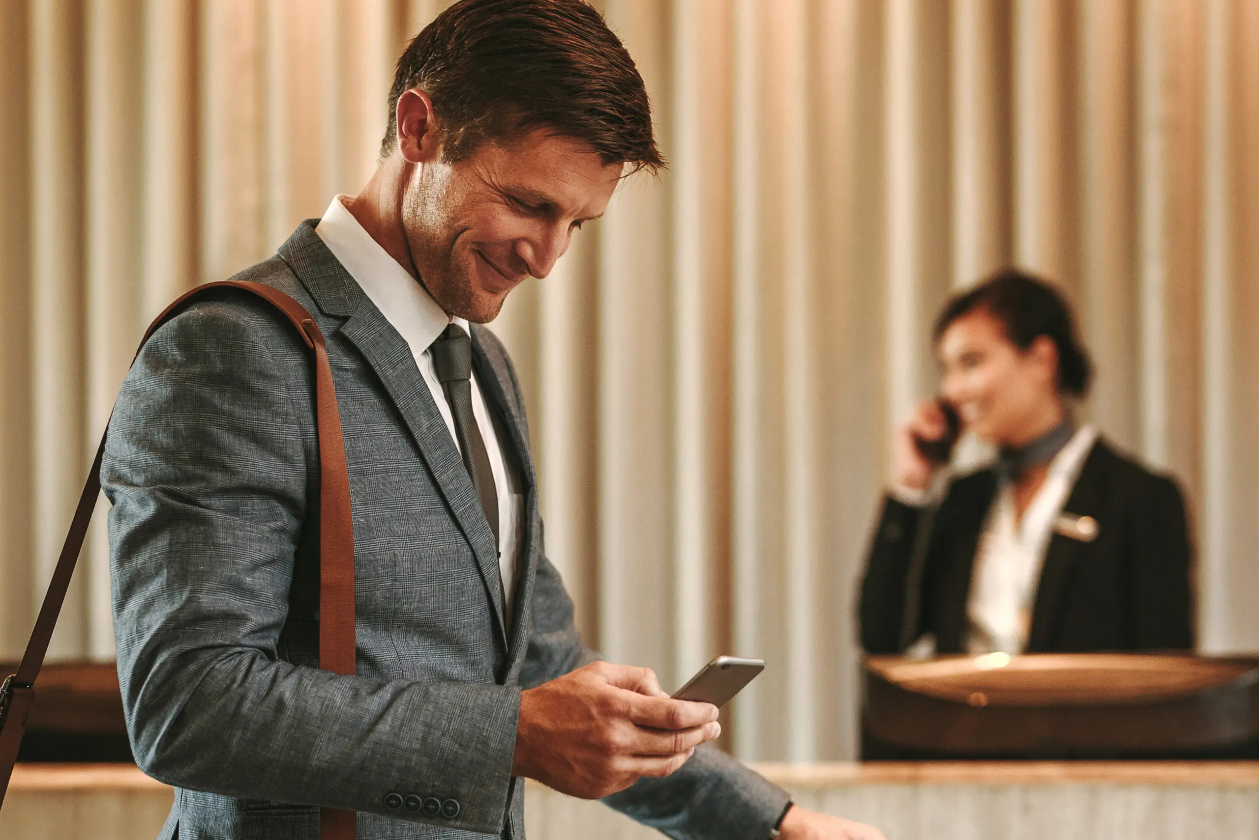 Man in a suit smiling at his phone, with a shoulder bag, standing near a hotel reception; a receptionist on a phone call in the background.