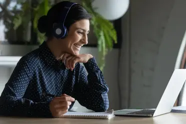 A woman wearing headphones smiles while looking at a laptop screen, sitting at a desk with a notepad.