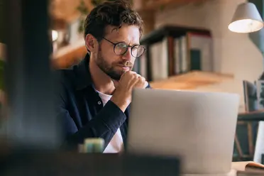 A man with glasses intently works on a laptop in a cozy, book-filled space with warm lighting.
