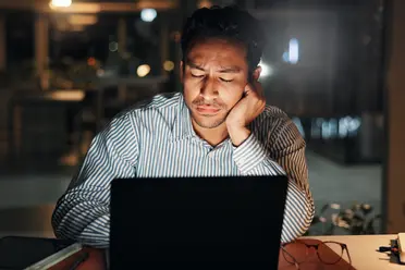 Man in a striped shirt looking tired, working on a laptop at night. Office setting with dim lighting in the background.