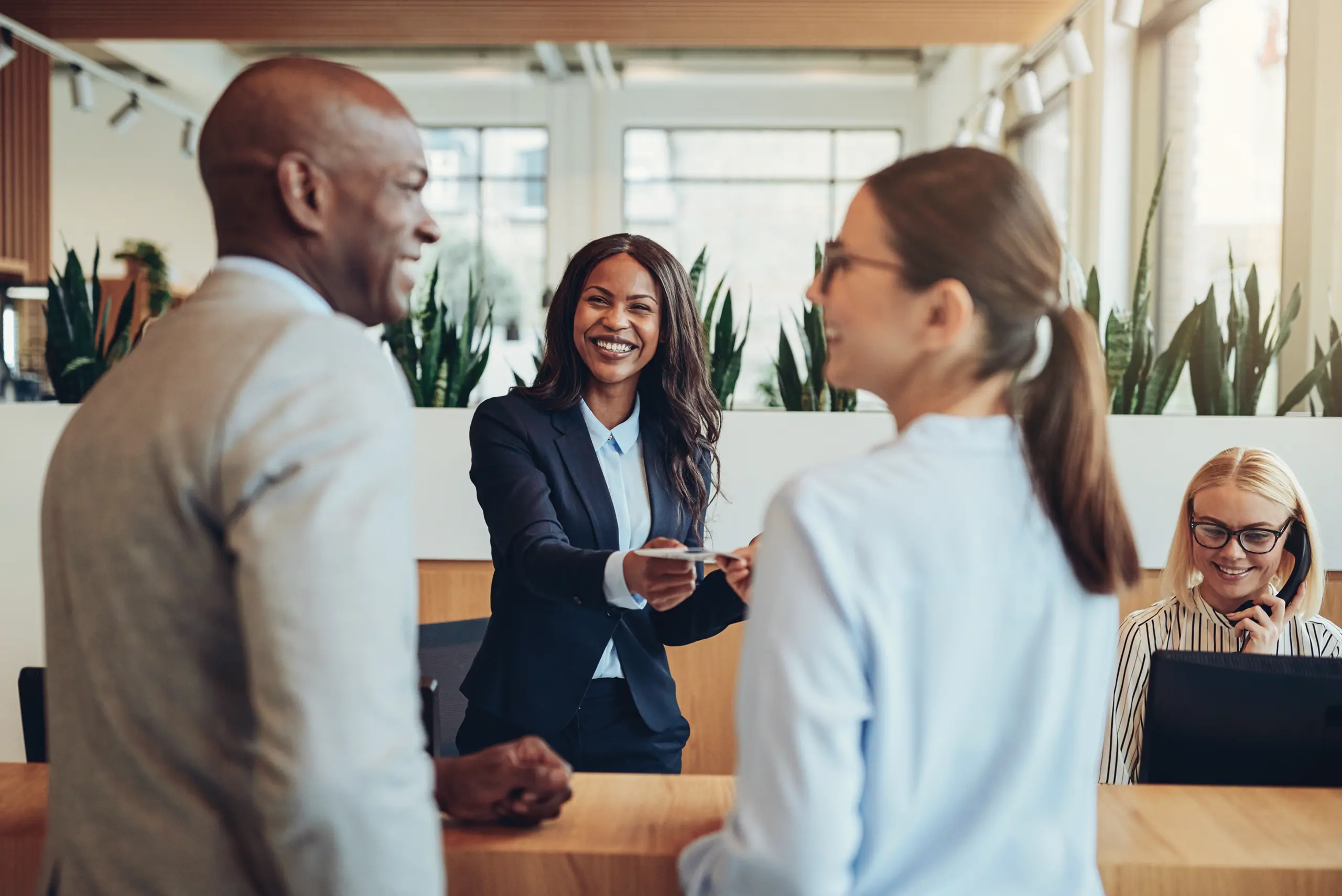 Businesspeople smiling and interacting at a reception desk, with one woman handing a document to a colleague in a bright, modern office setting.