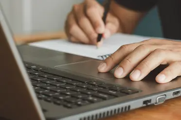 Close-up of a person's hand using a laptop touchpad, while the other hand writes on paper with a pencil.