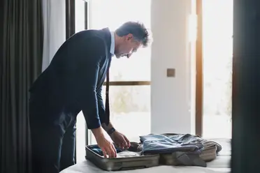 Man in a suit packing a suitcase on a bed in a bright room, with sunlight streaming through a nearby window.