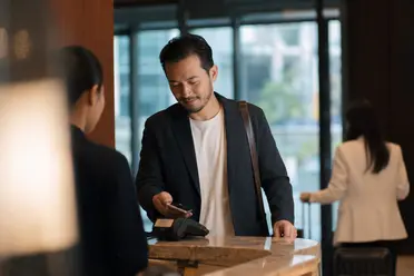 A man in a suit uses his phone at a hotel reception, while a woman with a suitcase approaches in the background.