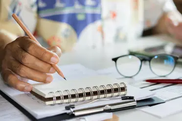 A person writing in a spiral notebook with a pencil, glasses nearby, and papers scattered on a desk.