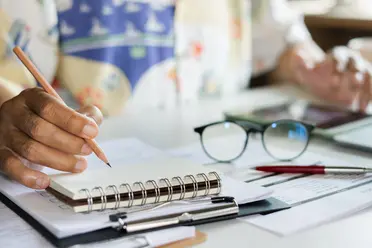 Person writing in a notebook at a cluttered desk with eyeglasses, pens, and papers scattered around.