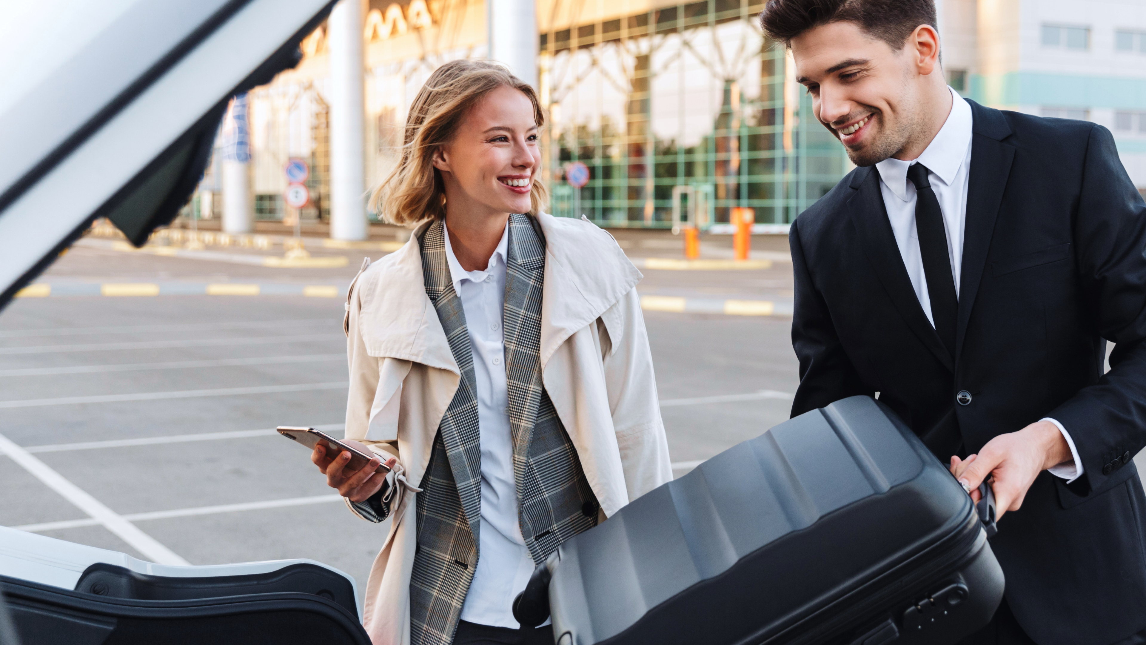 man loading suitcase into car boot while woman looks on, outside an airport