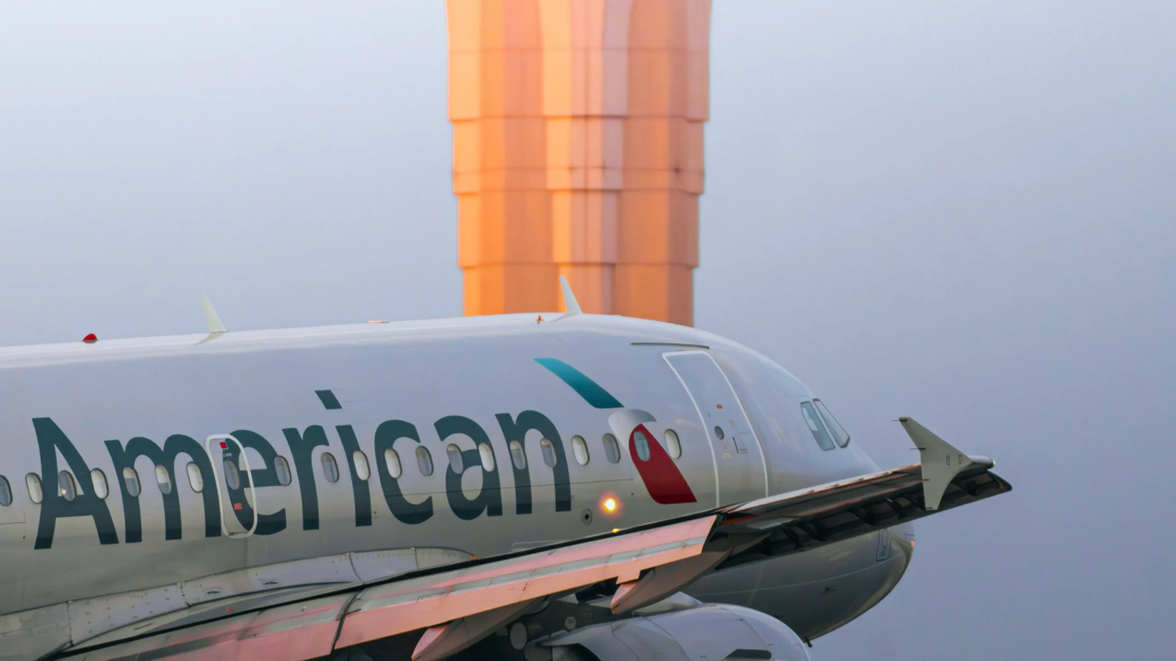 Close-up of American Airlines jet nose and cockpit with large orange airport control tower blurred in background.