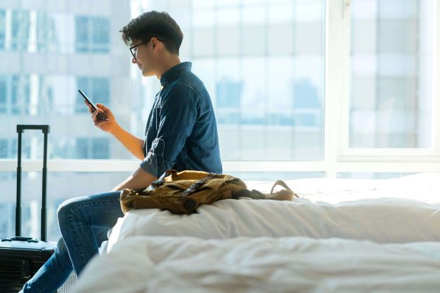 Mann sitz auf einem Hotelbett und schaut aufs Smartphone. Sein Rucksack liegt neben ihm auf dem Bett.