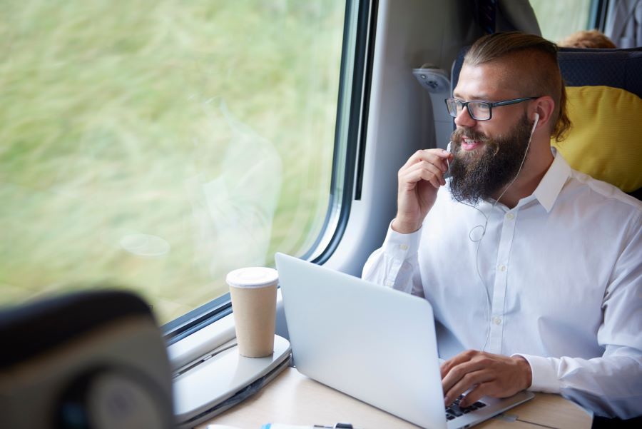 Mann im Zug sitzt an seinem Laptop und spricht in seine Kopfhörer