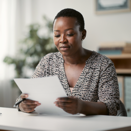 A Black British woman at a sunlit desk reading a single sheet of paperwork.