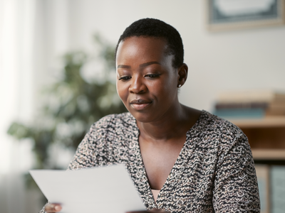 A Black British woman at a sunlit desk reading a single sheet of paperwork.