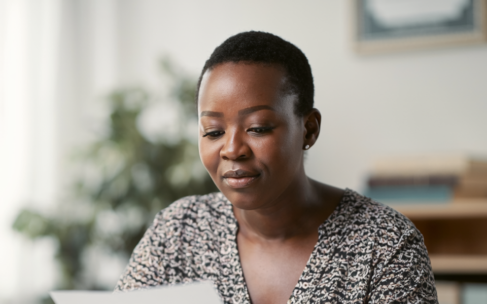 A Black British woman at a sunlit desk reading a single sheet of paperwork.