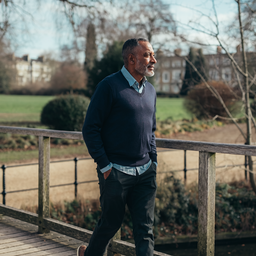 Man in his late forties walking across a wooden footbridge in a London park on a clear morning.