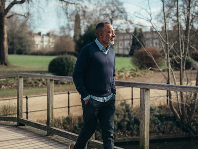 Man in his late forties walking across a wooden footbridge in a London park on a clear morning.