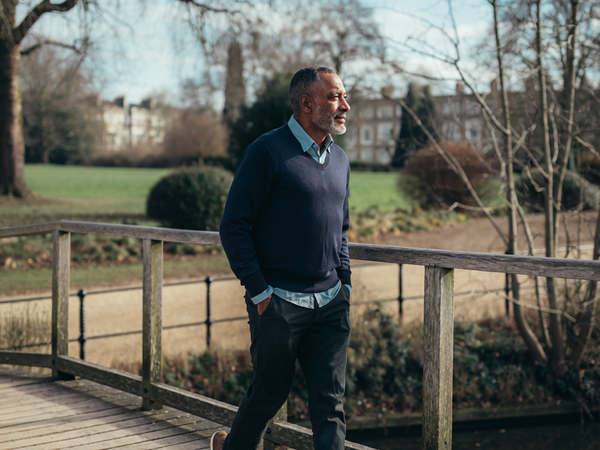 Man in his late forties walking across a wooden footbridge in a London park on a clear morning.