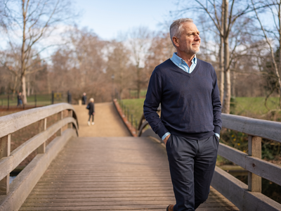 Man in his late forties walking across a wooden footbridge in a London park on a clear morning.