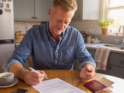 A man in his forties at a sunlit kitchen table filling in a visa application beside an open UK passport.