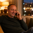 A man on a phone call at home, relaxed, in evening lamplight.
