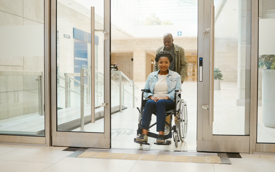 Wheelchair user and companion entering a bright, step-free healthcare area.