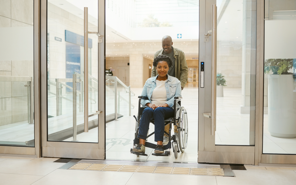 Wheelchair user and companion entering a bright, step-free healthcare area.