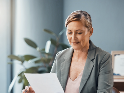 A woman in her fifties at a sunlit desk reading a single sheet of paperwork.