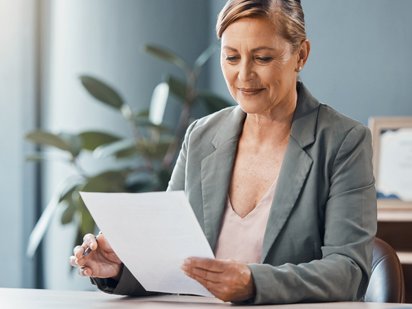 A woman in her fifties at a sunlit desk reading a single sheet of paperwork.