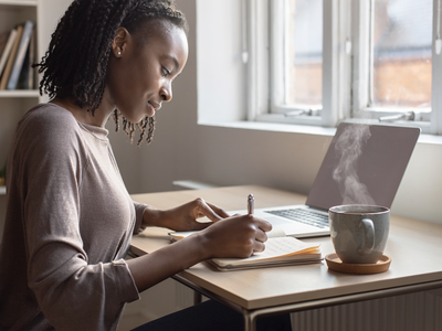 Black woman at home, focused on a notebook in natural daylight.