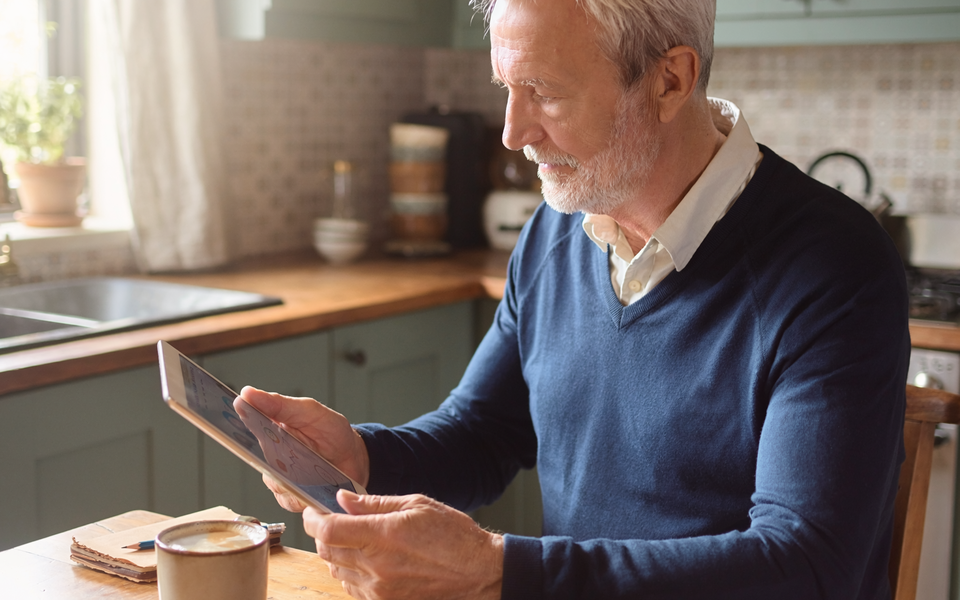 A man in his sixties reading a wellness chart on a tablet at a sunlit kitchen table.