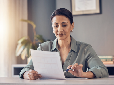 A South Asian woman at a sunlit desk reading a single sheet of paperwork.
