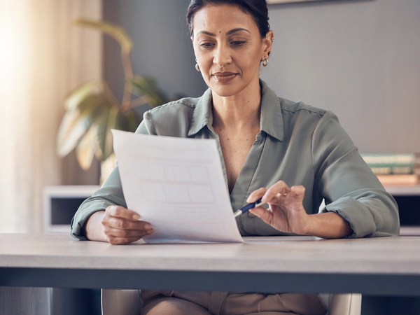 A South Asian woman at a sunlit desk reading a single sheet of paperwork.