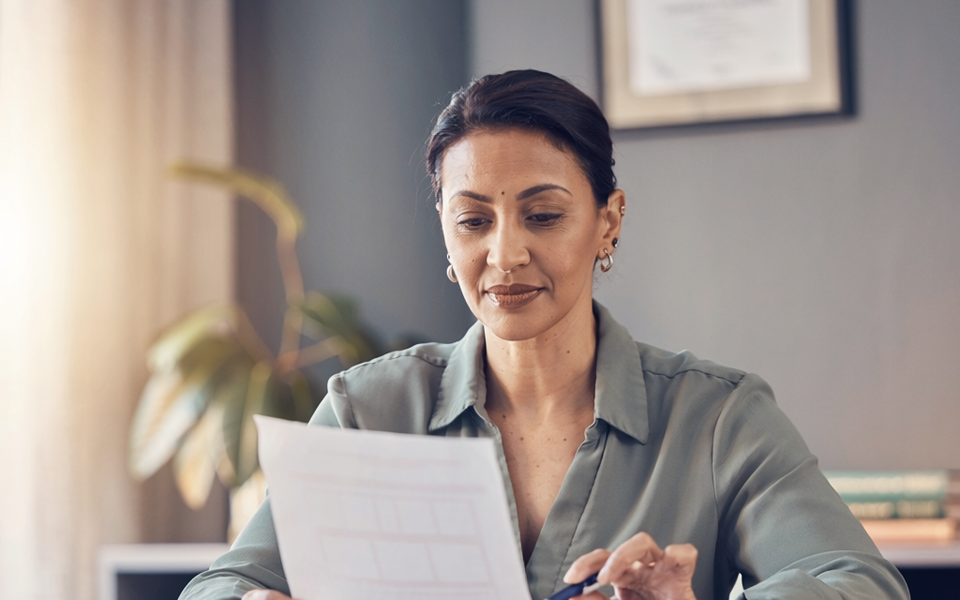 A South Asian woman at a sunlit desk reading a single sheet of paperwork.