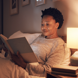 A woman in her 40s awake and calm in a dim bedroom at night, reading softly, not distressed