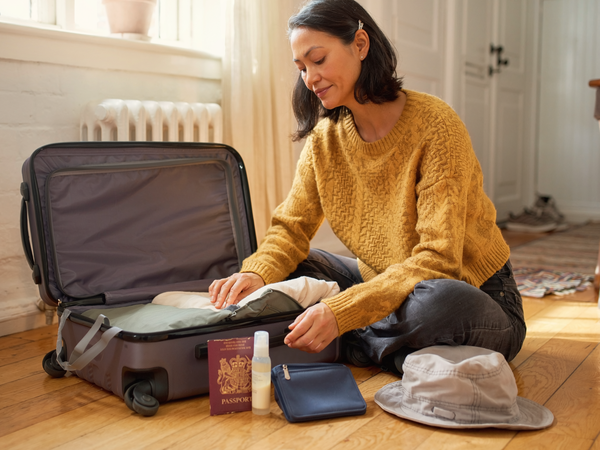 An East Asian woman packing a suitcase beside an open UK passport and travel essentials.