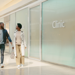 Two Londoners walking past a clinic frontage inside Westfield London on a bright weekday morning.