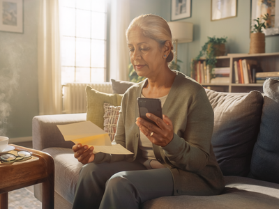A South Asian woman in her fifties at home, reading a paper letter and holding a smartphone.