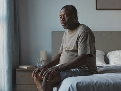 Black man in his fifties sitting on the edge of a bed in a calm London bedroom in the morning.