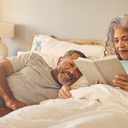 A couple in their 50s in bed in the morning, one waking gently, warm and undramatic