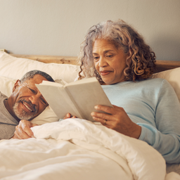 A couple in their 50s in bed in the morning, one waking gently, warm and undramatic
