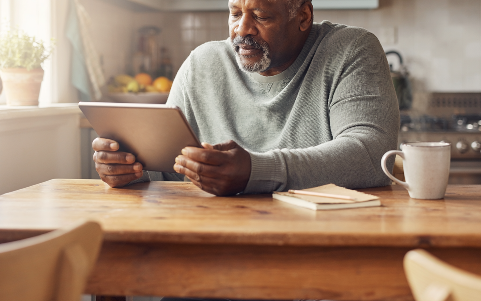 A Black British man in his fifties reading a wellness chart on a tablet at a sunlit kitchen table.