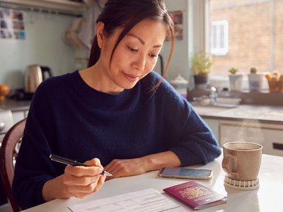 An East Asian woman at a sunlit kitchen table filling in a visa application beside an open UK passport.