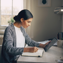South Asian adult at home, focused on a notebook in natural daylight.