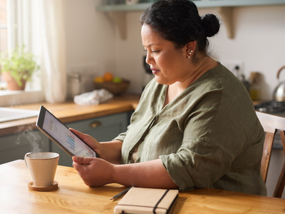 A South Asian woman in her forties reading a wellness chart on a tablet at a sunlit kitchen table.