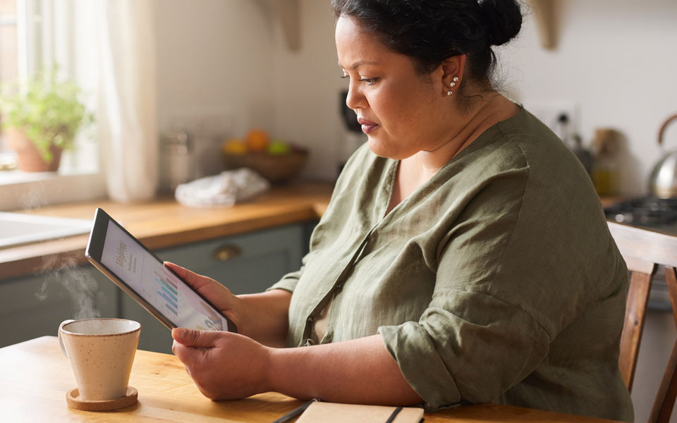A South Asian woman in her forties reading a wellness chart on a tablet at a sunlit kitchen table.