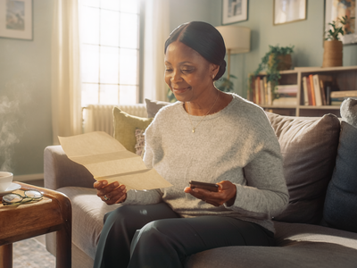 A Black British woman in her fifties at home, reading a paper letter and holding a smartphone.