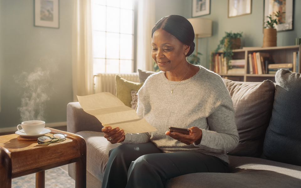 A Black British woman in her fifties at home, reading a paper letter and holding a smartphone.