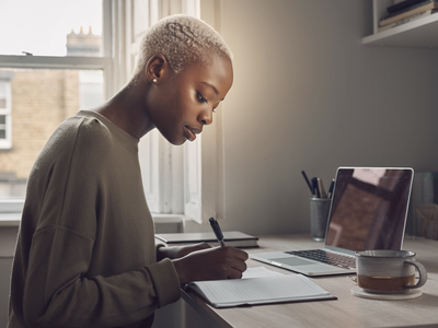 Black woman at home, focused on a notebook in natural daylight.